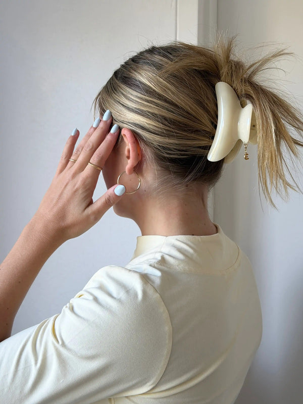 A woman shown from the back wearing a cream coloured, claw clip to hold up her blonde highlighted hair. She is wearing a cream coloured t-shirt and light blue nail polish, standing against a plain off white wall.