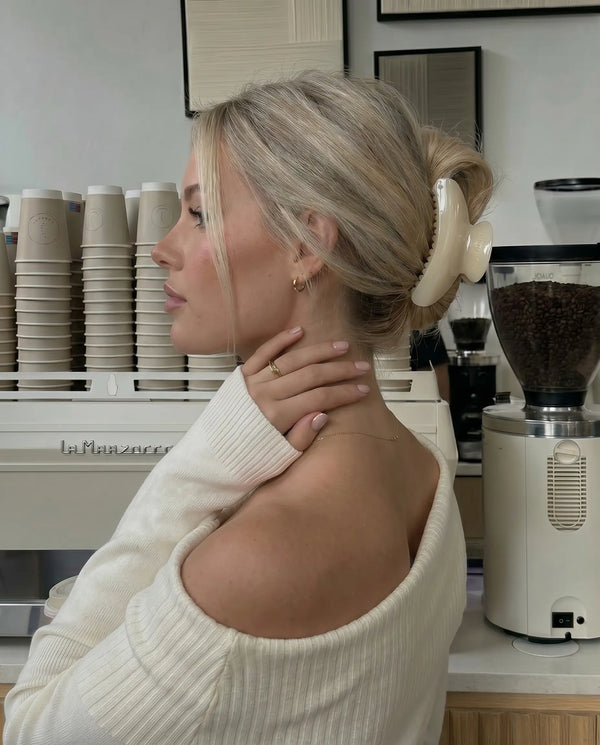 Woman in a white sweater sitting in a coffee shop with a coffee grinder and stacked cups in the background wearing a ivory coloured claw clip.