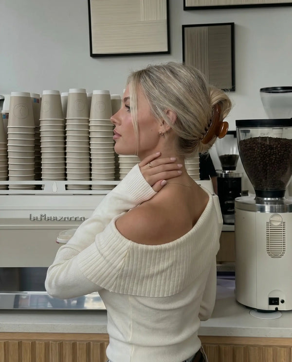 Woman in a white sweater standing in a coffee shop with coffee making equipment. Wearing a rich brown claw clip.