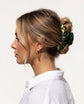 A side-profile view of a woman with blonde hair styled in a low bun, wearing a dark green and gold patterned hair claw clip and gold hoop earrings against a plain white background.
