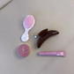 A styled flat-lay of beauty and hair accessories on a beige salon chair, including a pink and white hairbrush, a brown slim claw clip, a pink YSL compact mirror, and a pink lip balm tube.