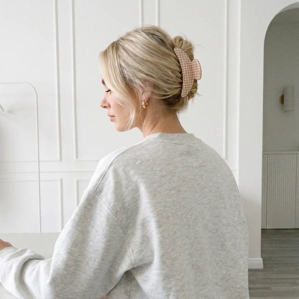 A woman with blonde hair in a messy bun secured with a slim pink and beige gingham hair claw clip, wearing a light gray sweatshirt in a bright white room.
