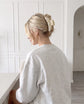 A blonde woman seen from behind wearing a light gray heather sweatshirt and a cream tiled coloured  hair claw clip in a bright, white panelled room.